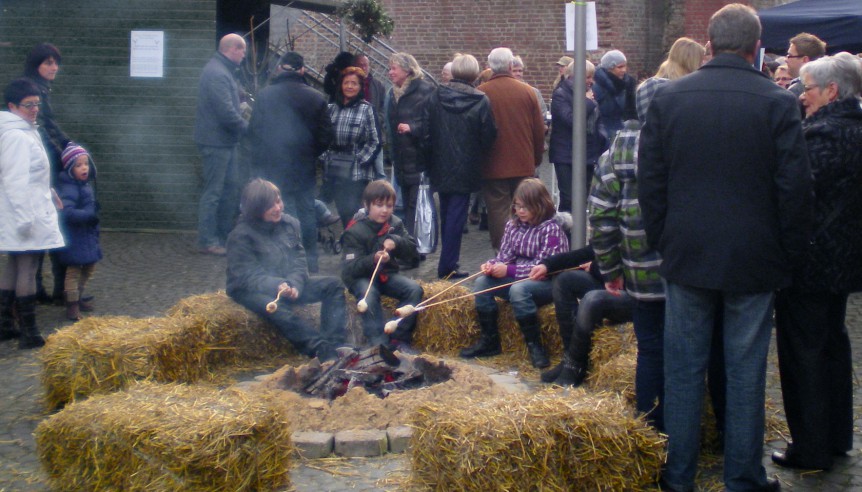 Weihnachtsmarkt vor der Burg Brüggen. Kinder sitzen auf Strohballen um ein Lagerfeuer und halten Stockbrot ins Feuer. Rundherum stehen Menschen, die sich unterhalten.
